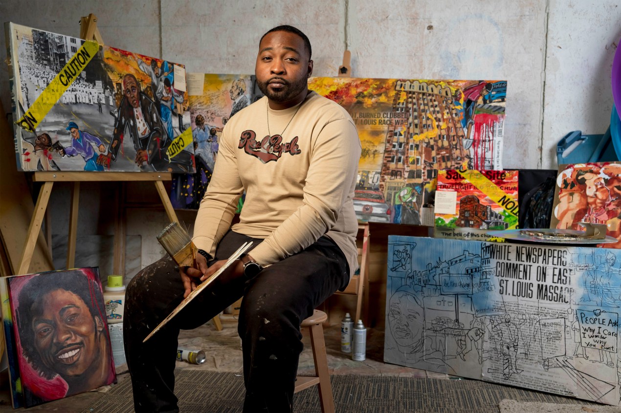 A photo shows Mykael Ash sitting in his studio, surrounded by his paintings behind him. He's holding a paintbrush and palette in his hands.