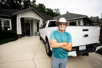 A man with his arms crossed in leans against a white pickup truck parked in a driveway.
