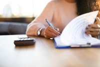 A person sits at a table with a phone, calculator and writes on paperwork on a clipboard.