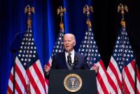 A photo of President Biden speaking at a podium with four American flags behind him.