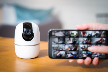 A photo of a small video camera device resting on a table. A hand holds up a phone screen in front of it, showing that camera feed can be monitored on the phone.